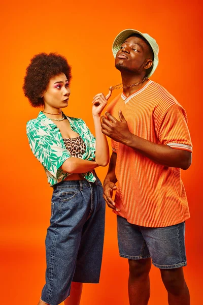 Escéptica mujer afroamericana con audaz maquillaje tocando la cadena en el mejor amigo en sombrero de panama y traje de verano aislado en naranja, amigos mostrando estilo individual, concepto de amistad - foto de stock