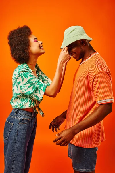 Vue latérale de la jeune femme afro-américaine souriante en tenue d'été touchant le chapeau panama sur le meilleur ami et s'amusant ensemble tout en se tenant isolé sur orange, amis en vêtements à la mode — Photo de stock