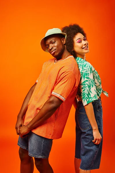 Молодой африканский американец в летней одежде и в панамской шляпе смотрит в камеру и стоит спиной к спине с лучшим другом с смелым макияжем и позирует изолированным на оранжевом, друзья в модной одежде — стоковое фото