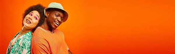 Young and pleased african american man in panama hat and summer outfit standing back to back with best friend with bold makeup isolated on orange, banner, friends in trendy clothes, friendship — Stock Photo