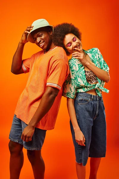 Jóvenes y alegres mejores amigos afroamericanos en trajes de verano elegantes posando mientras están de pie uno al lado del otro y pasar tiempo aislados en naranja, amigos en ropa de moda - foto de stock
