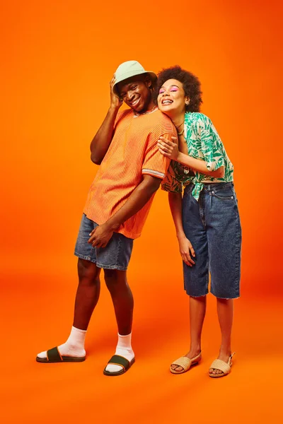 Longitud completa de sonriente joven afroamericana en traje de verano posando con el mejor amigo con estilo en sombrero de panama mientras está de pie sobre fondo naranja, amigos en ropa de moda, amistad - foto de stock