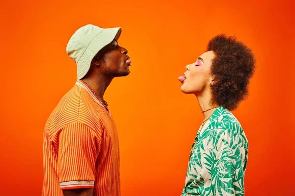 Side view of young and trendy african american best friends in summer outfits sticout tongues while standing together isolated on orange, friends in trendy clothes, friendship concept — стоковое фото