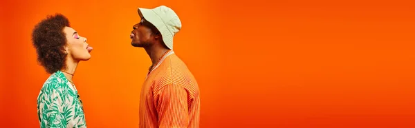 Side view of young and fashionable african american best friends in summer outfits sticking out tongues while standing together isolated on orange, banner, friends in trendy clothes — Stock Photo