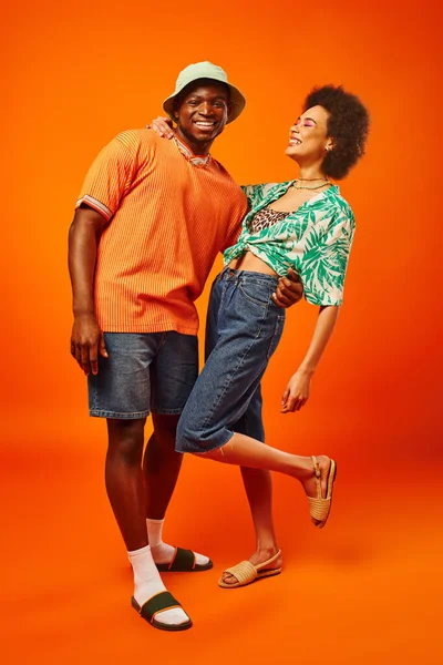 Comprimento total de jovem homem americano africano na moda em chapéu panama abraçando o melhor amigo satisfeito em roupa de verão, enquanto passa o tempo em fundo laranja, amigos em roupas da moda, amizade — Fotografia de Stock