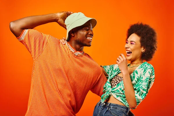 Jovem homem americano africano alegre em chapéu de panamá abraçando melhor amigo com maquiagem ousada e roupa de verão e se divertindo juntos isolados em laranja, amigos em roupas da moda, amizade — Fotografia de Stock