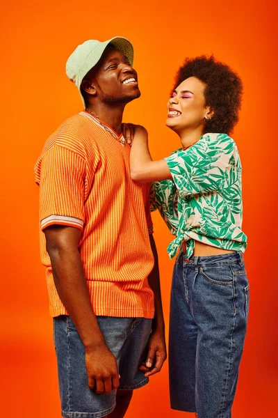 Mujer afroamericana joven positiva con maquillaje audaz y traje de verano abrazando al mejor amigo elegante en sombrero de panama mientras están de pie juntos aislados en naranja, amigos en ropa de moda - foto de stock