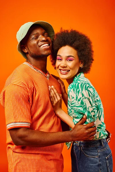 Portrait of pleased young african american man in panama hat and summer clothes hugging best friend with bold makeup and standing isolated on orange, friends in trendy clothes, friendship — Stock Photo