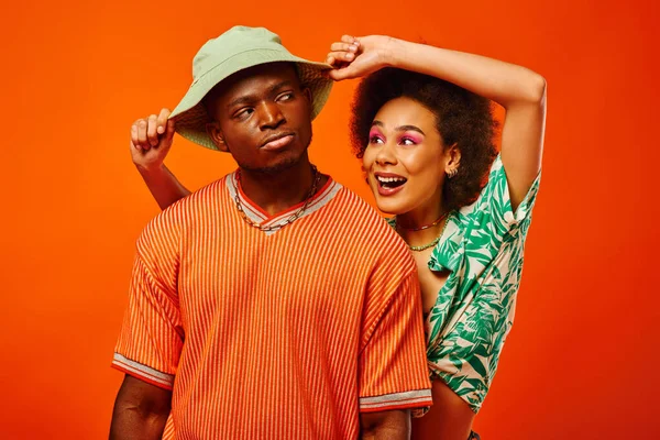 Smiling young african american woman with bold makeup in summer clothes touching panama hat of serious best friend and standing isolated on orange, friends in trendy clothes, friendship — Stock Photo