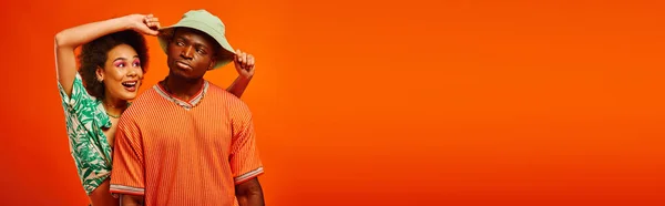 Positive junge afrikanisch-amerikanische Frau mit kühnem Make-up und sommerlichem Outfit, das Panamahut einer ernsthaften besten Freundin berührt, isoliert auf orangefarbenem Banner, modebewusste Freunde — Stock Photo