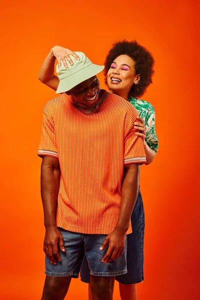 Позитивная молодая африканская американка с смелым макияжем трогает модного лучшего друга в панамской шляпе, позируя вместе изолированные на оранжевых, модных вперед друзей, дружбы — стоковое фото