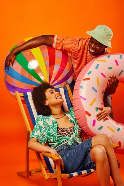 Positivo jovem afro-americano melhores amigos na roupa de verão elegante segurando bola de piscina e anel perto da cadeira de praia em fundo laranja, amigos da moda-frente, conceito de amizade — Fotografia de Stock