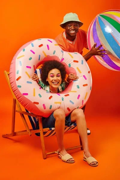 Positive young african american best friends in summer outfits holding pool ring and ball while sitting on deck chair and looking at camera on orange background, fashion-forward friends — Stock Photo