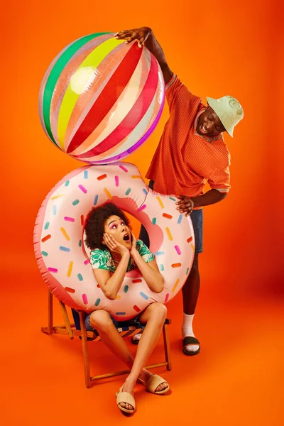 Positivo joven afroamericano hombre en traje de verano de moda celebración de la bola de bool cerca sorprendido mejor amigo con anillo de piscina en la silla de cubierta sobre fondo naranja, amigos de moda hacia adelante, amistad - foto de stock