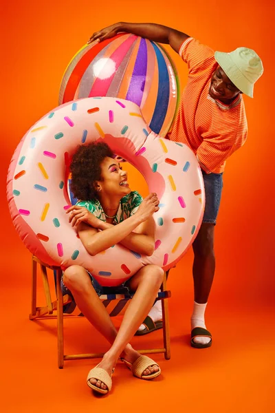 Jovem mulher americana africana alegre em roupa de verão segurando anel de piscina enquanto sentado na cadeira de praia e olhando para o melhor amigo com bola no fundo laranja, amigos da moda-forward — Fotografia de Stock