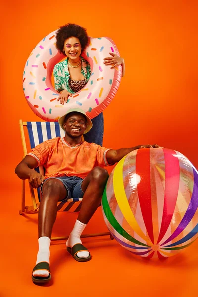 Full length of positive african american woman holding swim ring while standing near best friend in panama hat and summer outfit sitting on deck chair on orange background, fashion-forward friends — Stock Photo