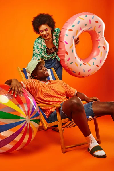 Smiling young african american woman in summer outfit holding swim ring while standing near stylish friend in panama hat sitting on deck chair on orange background, fashion-forward friends — Stock Photo