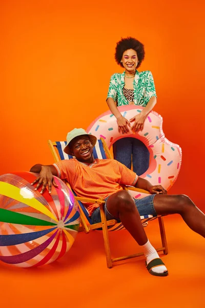 Amigos afroamericanos jóvenes positivos en trajes de verano que sostienen el anillo de natación y la bola cerca de la silla de cubierta mientras pasan tiempo juntos en el fondo naranja, amigos de moda hacia adelante, amistad - foto de stock