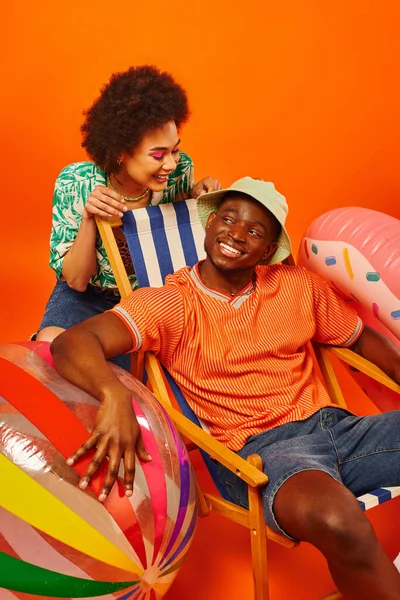 Радостная молодая африканская американка с смелым макияжем и летней одеждой, смотрящая на лучшего друга в панамской шляпе, сидящего на шезлонге рядом с мячом и плавающего кольца на оранжевом фоне, модных вперед друзей — стоковое фото