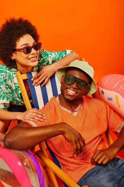 Sonrientes jóvenes afroamericanos mejores amigos en gafas de sol y trajes de verano pasar tiempo juntos cerca de la silla de cubierta, pelota de playa y anillo de natación sobre fondo naranja, amigos de moda hacia adelante - foto de stock