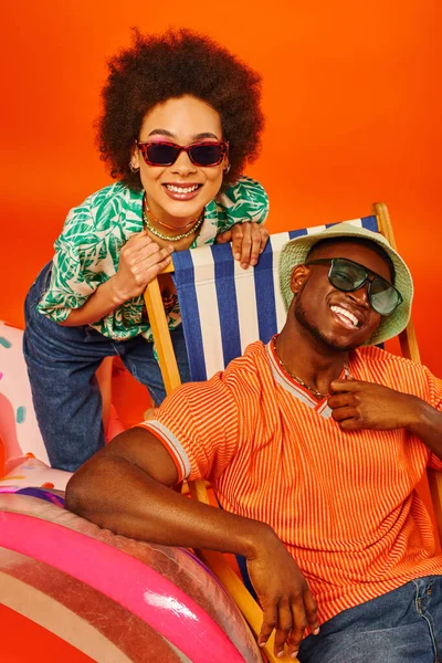 Mujer afroamericana joven positiva en gafas de sol y traje de verano de pie cerca de mejor amigo en sombrero de panama en la silla de cubierta y pelota de playa sobre fondo naranja, amigos de moda hacia adelante - foto de stock