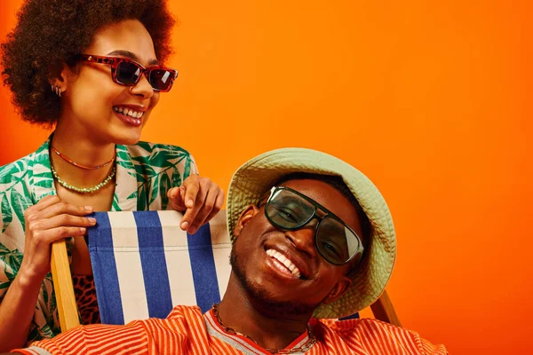 Mujer afroamericana joven positiva en gafas de sol y traje de verano de pie cerca de mejor amigo en sombrero de panama sentado en la silla de cubierta aislado en naranja, amigos de moda hacia adelante, amistad - foto de stock