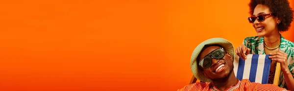Улыбчивая и стильная молодая африканская женщина в солнцезащитных очках и летнем костюме, стоящая рядом с лучшим другом в панамской шляпе, сидящая на шезлонге, изолированная на оранжевом, модные вперед друзья, баннер — стоковое фото