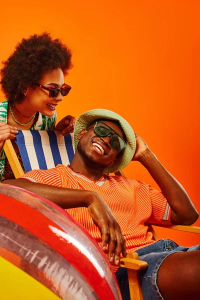 Pleased young african american woman in sunglasses and stylish summer outfit standing near best friend in panama hat relaxing on deck chair isolated on orange, friends in trendy casual attire — Stock Photo