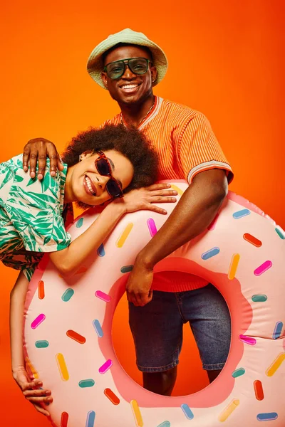 Joven afroamericano positivo con sombrero de panama y gafas de sol abrazando al mejor amigo de moda en traje de verano y sosteniendo un anillo de baño aislado en naranja, amigos con atuendo casual de moda - foto de stock