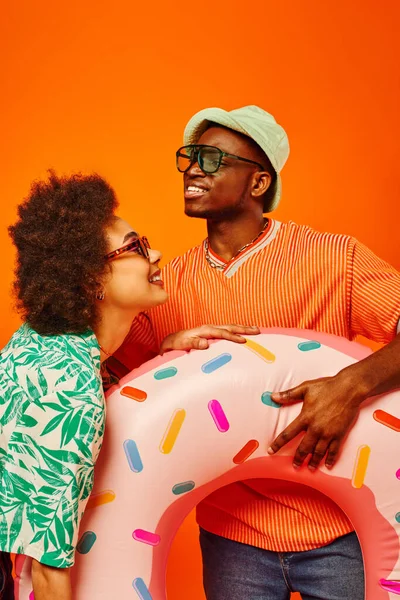 Jovens amigos afro-americanos satisfeitos em óculos de sol e roupas de verão segurando anel de natação, passando o tempo e de pé juntos isolados em laranja, amigos em trajes casuais na moda — Fotografia de Stock