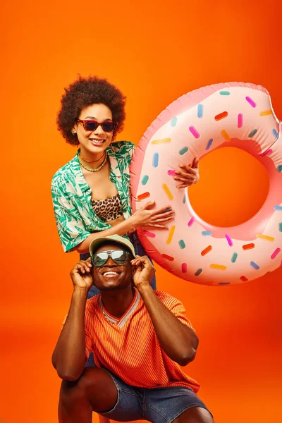 Jovem homem americano africano alegre em óculos de sol e chapéu de panamá posando perto melhor amigo na roupa de verão na moda segurando anel de natação isolado em laranja, amigos em trajes casuais na moda — Fotografia de Stock