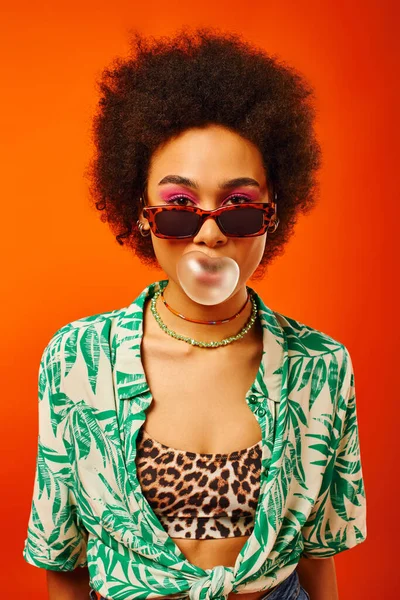 Портрет молодой и модной африканской женщины с смелым макияжем в солнцезащитных очках и летнем наряде и жвачкой, изолированной на красном, женщина с чувством стиля, уверенностью — стоковое фото