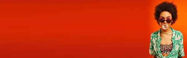 Mujer afroamericana joven con maquillaje audaz que usa traje de verano y gafas de sol mientras sopla goma de mascar mientras está de pie aislado en rojo, mujer con sentido del estilo, bandera, confianza - foto de stock