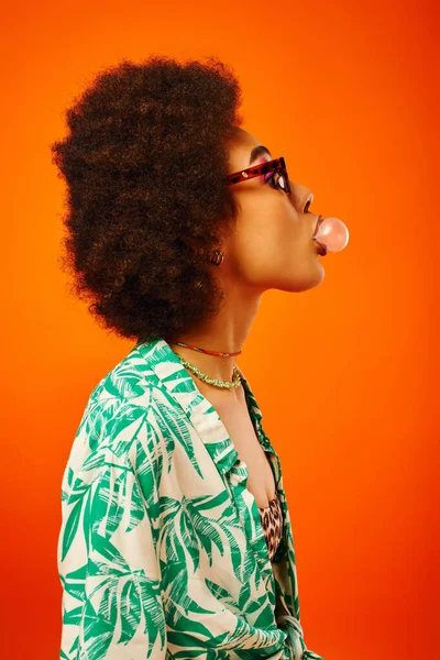 Vue latérale de jeune femme afro-américaine à la mode portant des lunettes de soleil en tenue d'été tout en soufflant de la gomme à bulles et se tenant isolée sur le rouge, femme avec un sens du style, confiance — Photo de stock