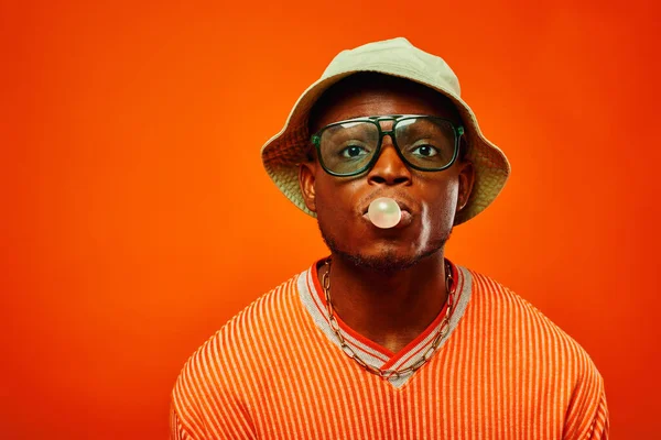 Portrait of young and stylish african american man in summer outfit wearing sunglasses and panama hat blowing bubble gum and looking at camera isolated on red, man with sense of style — Stock Photo