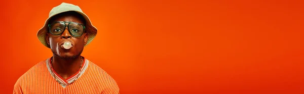 Giovane afroamericano alla moda in abito estivo, occhiali da sole e cappello panama che soffia gomma da masticare e guardando la fotocamera isolata sul rosso, uomo con senso dello stile, banner, fiducia — Foto stock