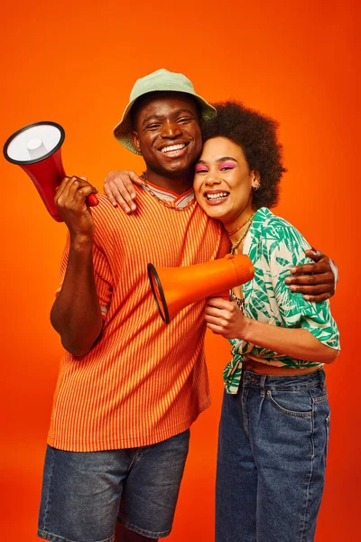 Portrait of cheerful young african american man in summer outfit and panama hat holding loudspeaker and hugging best friend with bold makeup isolated on red, friends in fashionable outfits — Stock Photo