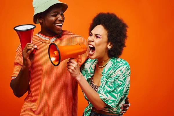 Cheerful young african american man in summer outfit and panama hat looking at stylish best friend screaming at loudspeaker and standing isolated on red, friends in fashionable outfits — Stock Photo