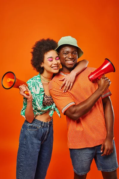 Улыбающаяся молодая африканская американка с смелым макияжем и летней одеждой обнимает лучшего друга в панамской шляпе и держит громкоговоритель, стоя изолированно на красном, друзей в модных нарядах — стоковое фото