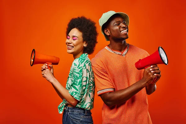 Cheerful young african american best friends in modern summer outfits holding loudspeakers and standing next to each other isolated on red, friends in fashionable outfits, friendship — Stock Photo
