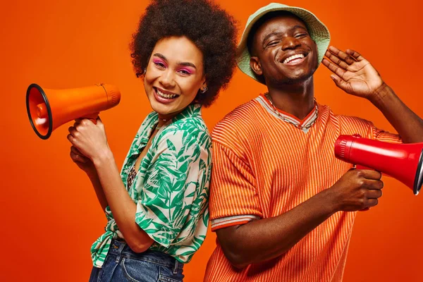 Positive young african american best friends in summer clothes looking at camera while holding loudspeakers and standing isolated on red, friends in fashionable outfits, friendship — Stock Photo