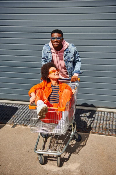 Smiling young african american man in denim jacket and sunglasses standing near best friend in shopping cart and building on urban street, friends hanging out together, friendship — Stock Photo