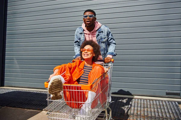 Jeune homme afro-américain excité avec des lunettes de soleil et une veste en denim debout près d'un meilleur ami souriant assis dans un chariot et construisant dans une rue urbaine, des amis traînant ensemble — Photo de stock