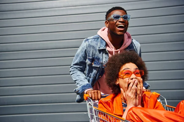 Cheerful young african american man in sunglasses having fun with scared best friend in trendy outfit and shopping cart near building on urban street, friends hanging out together — Stock Photo