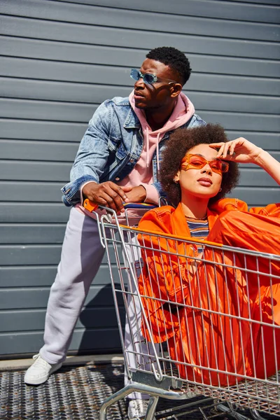 Elegante jovem afro-americana em roupa brilhante e óculos de sol olhando para a câmera enquanto sentado no carrinho de compras perto de um amigo da moda e construindo na rua urbana, amigos saindo juntos — Fotografia de Stock