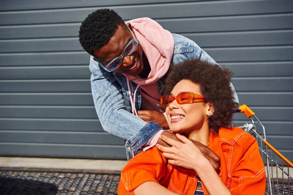 Pleased young african american man in denim jacket and sunglasses hugging trendy best friend sitting in shopping cart and spending time on urban street, friends with stylish vibe concept — Stock Photo