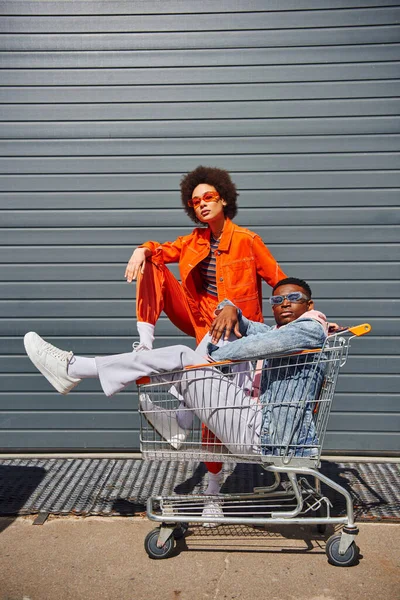 Full length of trendy young african american woman in sunglasses and bright outfit posing near best friend sitting in shopping cart and looking at camera on urban street, friends with stylish vibe — Stock Photo