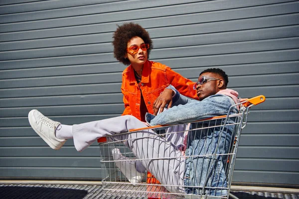 Stilvolle junge afrikanisch-amerikanische Frau mit Sonnenbrille und hellem Outfit, die in der Nähe des besten Freundes in Jeansjacke im Einkaufswagen sitzt und draußen baut, Freunde mit stilvollem Ambiente — Stockfoto