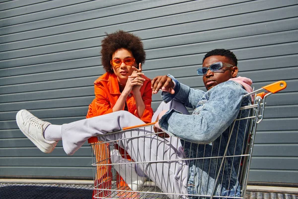 Selbstbewusst und trendy junge afrikanisch-amerikanische beste Freunde in Sonnenbrille und hellem Outfit posieren mit Einkaufswagen in der Nähe von Gebäuden an der Stadtstraße, Freunde mit stilvollem Vibe-Konzept — Stockfoto