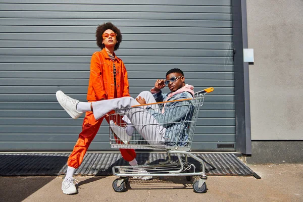 Full length of trendy and confident young african american best friends in sunglasses and bright outfits looking at camera and posing with shopping cart on urban street, friends with stylish vibe — Stock Photo